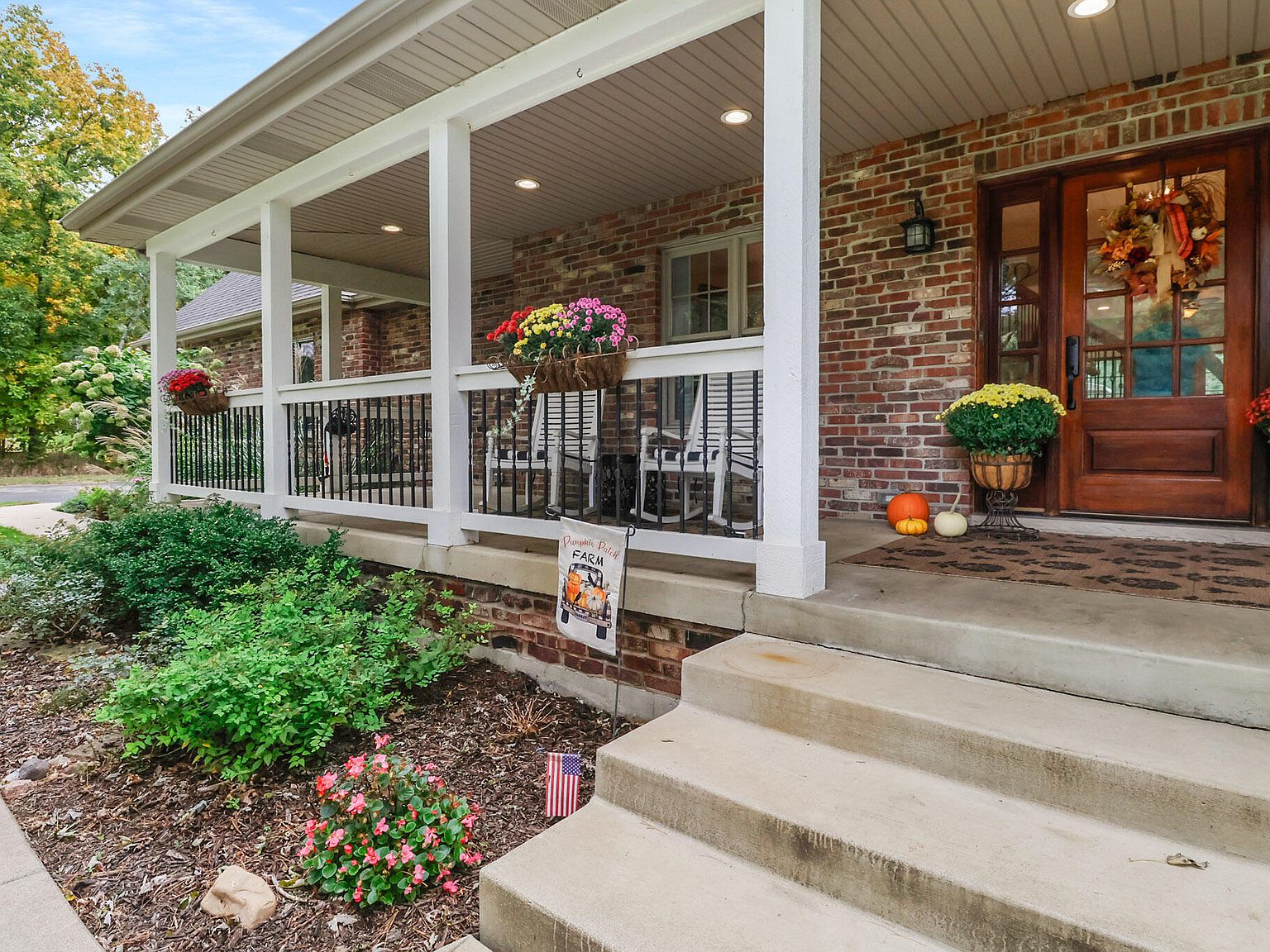 The covered front porch at 9905 W 113th Ave, with rocking chairs, fall mums in planters, an autumn wreath on the wooden front door, and brick columns framing the porch
