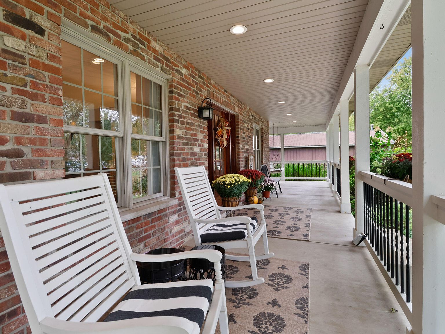 A long view down the wraparound porch with paired white rocking chairs on a patterned rug, brick wall on one side, dark iron spindles set into the wood porch rail on the other, and a glimpse of the rear pole barn through the porch posts