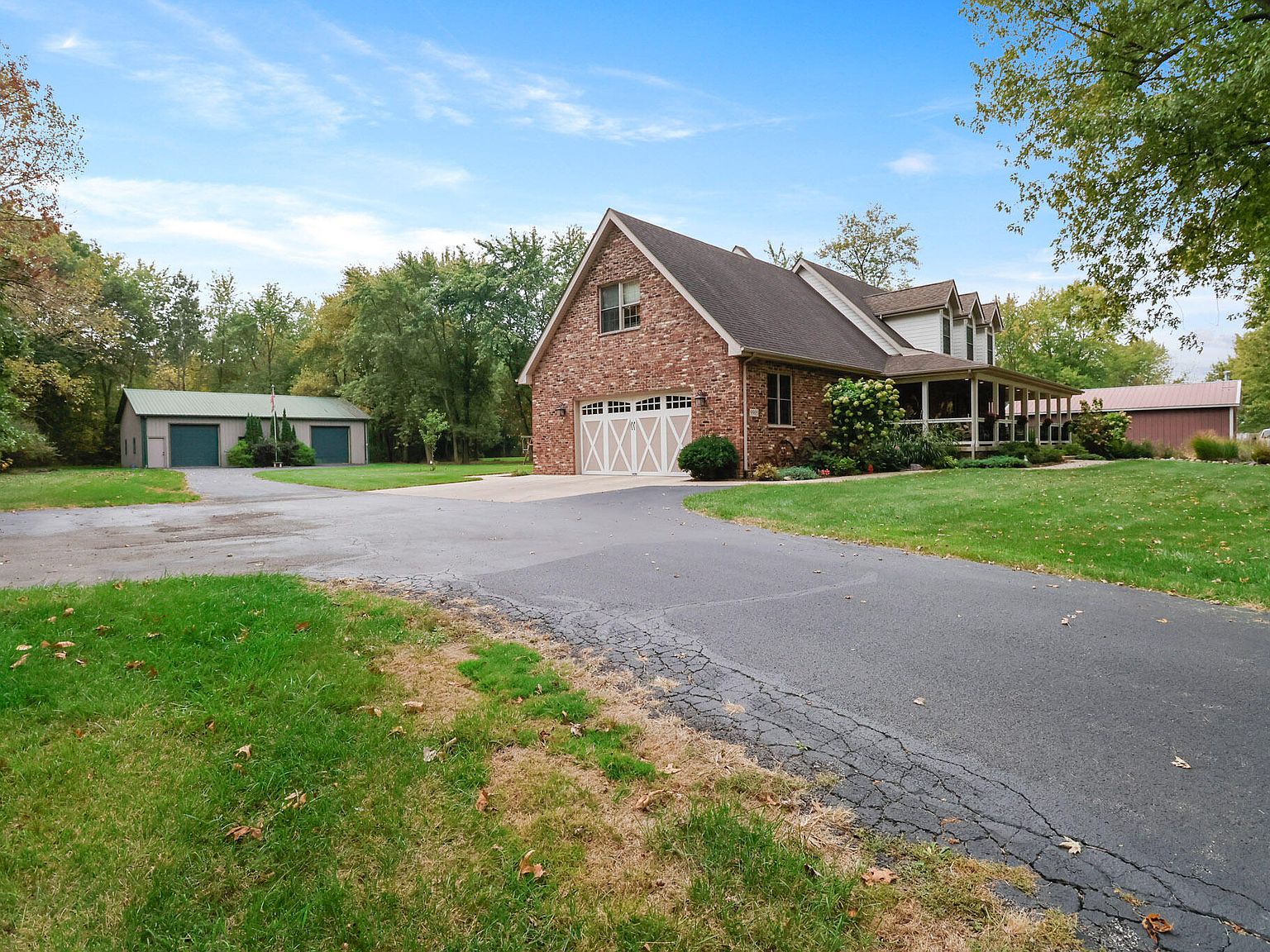 Side angle of the home showing the attached two-car garage on the brick side elevation, the detached white pole barn beyond a paved driveway, and the property's tree line behind both structures