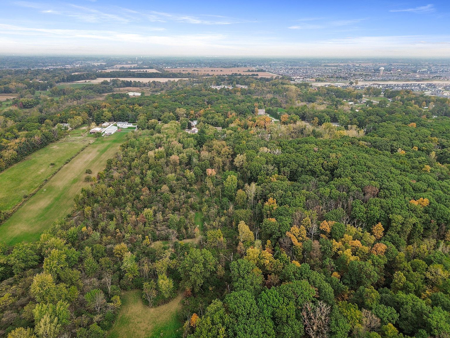 Aerial view of 9905 W 113th Ave showing 5.67 acres of dense mixed-hardwood canopy in autumn colors, the home's white roofline tucked into the trees at the left, with the surrounding cleared neighborhood and a distant town skyline visible on the horizon