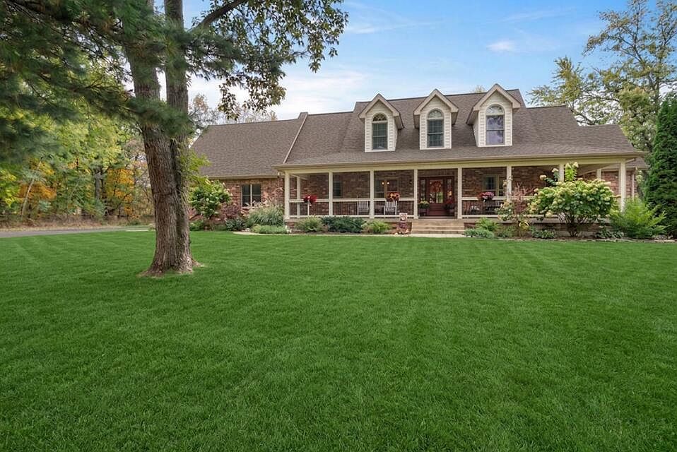 Wider front-elevation view of 9905 W 113th Ave across the manicured front lawn, the three dormers and wraparound porch in full view, the mature oak in the foreground anchoring the left of the frame